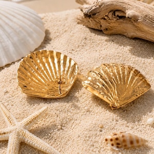 Pode incluir: Um par de brincos de metal dourado em forma de conchas de vieira. Os brincos apresentam uma superfície texturizada e canelada que imita as cristas naturais de uma concha, com um acabamento polido que reflete a luz sobre a forma curva.
