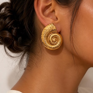 Könnte beinhalten: Ein goldfarbener Ohrring in Form eines Nautilus-Gehäuses mit polierter metallischer Oberfläche und komplexen, erhabenen Spiralrippen, die dem natürlichen Schwung des Muscheldesigns folgen und mit einem glatten, runden Verschluss am Ohrläppchen befestigt sind.