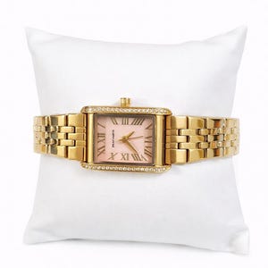 May include: A gold-coloured rectangular wristwatch with a pink face and Roman numeral hour markers. The watch has a gold link band and is decorated with small crystals around the face. The watch is displayed on a white cushion.