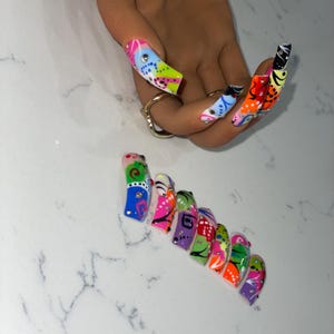 May include: A hand with long, colorful, and patterned artificial nails. The nails feature a variety of designs, including abstract shapes, dots, and lines, in vibrant colors like pink, blue, green, and orange. Some nails have small, sparkling embellishments.