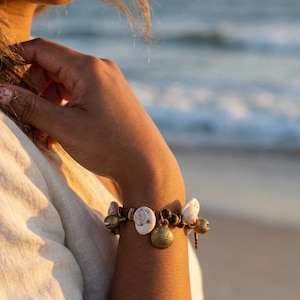 May include: A bracelet featuring a dark brown cord adorned with white spiral seashells, small dark brown beads, and several metallic gold-toned charms shaped like shells and small bells, secured with copper-colored wire wrapping.