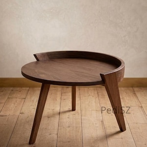 May include: A dark brown, round wooden side table with three angled legs. The table has a raised edge on the top surface and a cutout on one side. The table is set on a wooden floor.