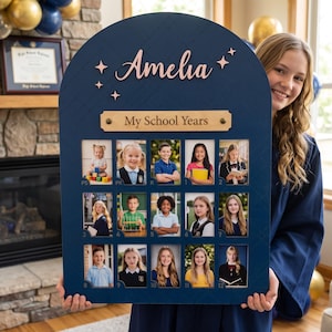 May include: A dark blue arched wooden school photo display board featuring the name Amelia in rose gold script, star accents, and a light wood plaque labelled My School Years. It contains fifteen rectangular slots for student portraits.