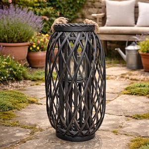 May include: A dark brown woven lantern with a cylindrical shape. The lantern has a rope handle and a glass insert for a candle. It is placed on a stone patio, with plants and a bench in the background. The lantern is approximately 61 cm tall.
