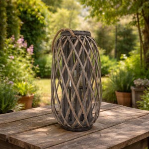 May include: A cylindrical woven lantern with a natural wood finish and a rope handle. The lantern has a lattice design. It is placed on a wooden table in a garden setting.