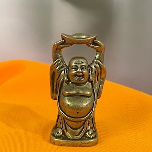 Könnte beinhalten: Kleine Buddha-Statue in Bronzefarbe mit lächelndem Gesicht und erhobenen Armen, die einen Goldbarren halten. Die Figur ist in sitzender Position dargestellt und trägt Gewänder. Die Statue steht auf einer orangefarbenen Oberfläche.