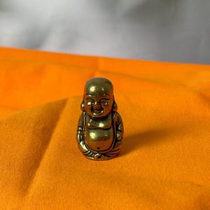 Puede incluir: Pequeña estatua de Buda de color latón con una expresión sonriente. La figura está representada en posición sentada, con una barriga redonda y rasgos faciales detallados. La estatua está sobre un fondo de tela naranja.