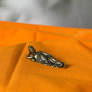 May include: A small, bronze-colored Buddha statue in a reclining pose. The figure is detailed with flowing robes and is resting on an orange fabric. The statue is a representation of Buddhist art and spirituality.