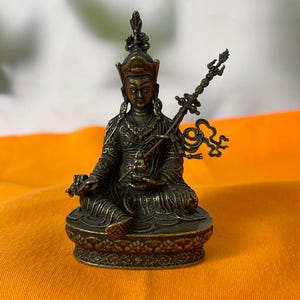 May include: A bronze-colored statue of a seated figure, adorned with a detailed headdress and ornate clothing. The figure holds a staff with intricate designs. The statue is placed on a decorative base, set against an orange and white background.