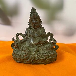 May include: A small, dark bronze-toned metal figurine depicting a seated deity with an ornate headdress, prominent belly, and detailed robes. The figure rests on a textured, circular base with intricate carvings and flowing elements.