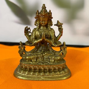 Könnte beinhalten: Eine kleine, goldfarbene Metallstatue einer vierarmigen Gottheit, die im Schneidersitz auf einem Lotussockel sitzt. Die Figur trägt eine verzierte Krone und Schmuck, wobei zwei Hände zum Gebet gefaltet sind und zwei Hände erhoben sind.
