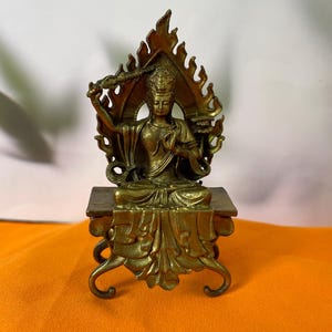 May include: A bronze-toned metal figurine of a seated deity with a raised right arm holding a staff, positioned before a flame-shaped mandorla. The figure rests on a rectangular pedestal with ornate, curved legs and draped fabric detailing.