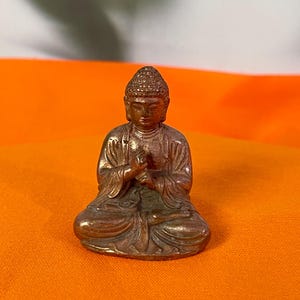 May include: A small bronze Buddha statue in a seated meditation pose. The statue has detailed features and is set against an orange background. The Buddha is wearing a robe and has a serene expression.