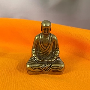 May include: A small, gold-colored statue of a seated figure in a meditative pose. The figure is wearing a draped robe and has a serene expression. The statue is on an orange fabric background.
