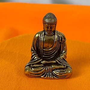 May include: A small, bronze-colored Buddha statue in a seated meditation pose. The figure has detailed robes and a serene expression. The statue is set against an orange background, highlighting its intricate design and spiritual theme.