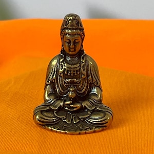 Puede incluir: Una pequeña estatua de bronce de un Buda sentado en posición meditativa. La figura tiene ropa detallada y una expresión serena. La estatua está sobre un fondo naranja.