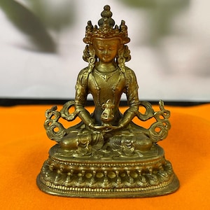 May include: A small, metallic gold-coloured statue of a seated deity. The figure wears an ornate crown and jewellery, with hands resting in a meditative pose holding a small vessel. It sits upon a detailed, tiered lotus-style pedestal base.