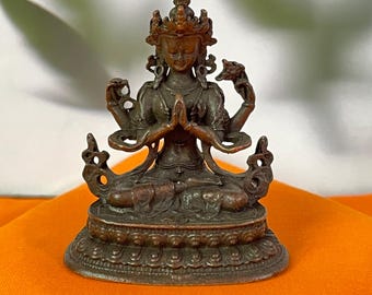 May include: A bronze-colored statue of a seated deity with multiple arms and a crown. The figure is in a meditative pose, with hands clasped in prayer. The statue is on a detailed circular base, set against an orange background.