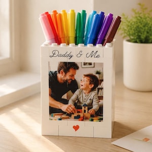 May include: A white pen holder with the words "Daddy & Me" and a photo of a father and child. The holder is filled with colourful markers. A small red heart is at the bottom of the holder.