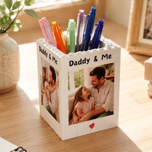 Könnte beinhalten: Ein weißer Stifthalter im Blockstil mit dem Aufdruck "Daddy & Me". Der Halter enthält mehrere Stifte in verschiedenen Farben. Der Halter hat zwei Familienfotos und ein rotes Herz. Im Hintergrund befindet sich eine Vase mit Grünzeug.