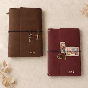May include: A brown leather journal with the initials L.M.N. and a burgundy leather journal with the initials JR and a heart symbol. Both journals feature black elastic bands with metal charms, including a key, aeroplane, anchor, and statue.