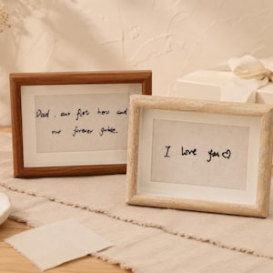Peut inclure: Deux cadres photo rectangulaires en bois avec passe-partout blanc. Le cadre de gauche a une finition en bois foncé et contient le texte "Dad, our first hero and our forever guide". Le cadre de droite a une finition en bois clair et indique "I love you" avec un cœur.
