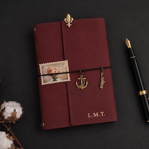 Peut inclure: Un carnet en cuir bordeaux profond avec une attache fleur de lys dorée sur le dessus, les initiales L.M.T. gravées en or, et un élastique noir retenant un timbre floral rectangulaire ainsi que deux breloques dorées en forme d'ancre et de Statue de la Liberté.
