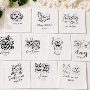 Op de afbeelding: Een verzameling witte papieren servetten met illustraties van honden en katten in zwarte inkt. Elke servet heeft een ander ontwerp, waaronder dieren met tekst als "Drinks on me!" en "Happy Birthday!"