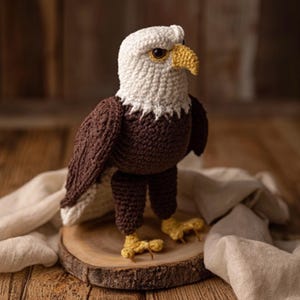 May include: A handmade crochet eagle with a white head, brown body, and yellow beak and feet. The eagle is standing on a wooden slice, showcasing its detailed craftsmanship. The bird's wings are spread, and it has a focused gaze.