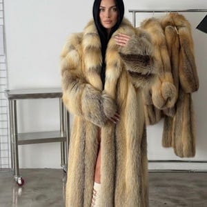 Puede incluir: Un abrigo largo y lujoso de piel en tonos beige, marrón y crema. El abrigo tiene una silueta amplia y lo lleva una persona. Abrigos de piel adicionales cuelgan de una percha en el fondo, lo que sugiere un tema de moda o ropa de abrigo.