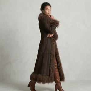 May include: A long, dark brown coat with a faux fur trim around the collar, cuffs, and hem. The coat is made of a suede-like material and has a tailored fit. The model is wearing brown leather boots.