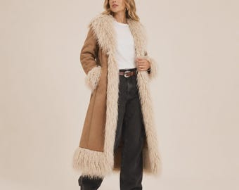 May include: A long tan coat featuring a thick, shaggy cream-colored faux fur trim along the collar, front opening, cuffs, and hem. The coat is worn over a plain white t-shirt and dark charcoal gray denim jeans with a brown leather belt.