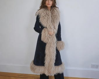 May include: A long, dark navy blue coat featuring a prominent, thick, beige-coloured curly fur trim along the wide collar, down the front opening, around the sleeve cuffs, and along the bottom hemline. The coat is worn over dark trousers and black shoes.
