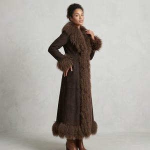 May include: A long, dark brown coat with a faux fur trim around the collar, cuffs, and hem. The coat is made of a suede-like material and has a tailored fit. The model is wearing brown boots.