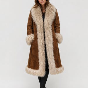 May include: A long brown coat with a cream-colored faux fur collar, cuffs, and trim. The coat is knee-length and has a classic, vintage style. The model is wearing black pants and black boots.