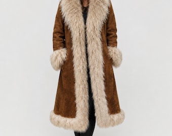 May include: A long brown coat with a cream-colored faux fur collar, cuffs, and trim. The coat is knee-length and has a classic, vintage style. The model is wearing black pants and black boots.