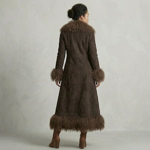 May include: A long, brown coat with a faux fur collar, cuffs, and hem. The coat is made of a suede-like material and has a tailored fit. The model is wearing brown boots. The coat is a stylish outerwear option.