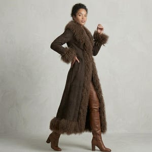 May include: A long, brown coat with a faux fur collar, cuffs, and hem. The coat is worn with brown leather pants and knee-high boots. The model is standing in front of a neutral background. The coat appears to be made of suede or a similar material.