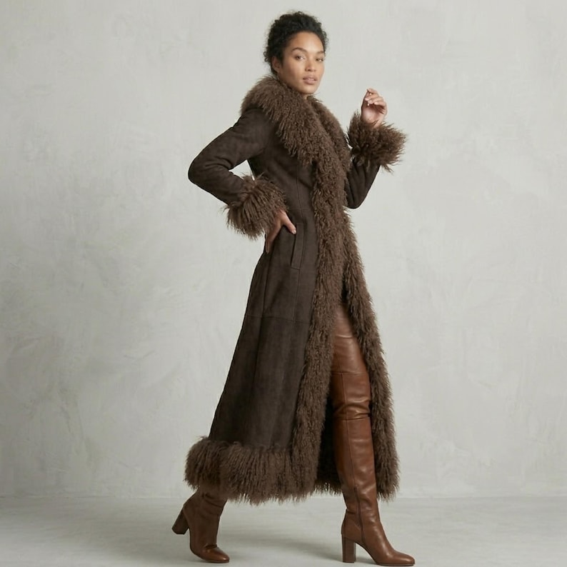 May include: A long, brown coat with a faux fur collar, cuffs, and hem. The coat is worn with brown leather pants and knee-high boots. The model is standing in front of a neutral background. The coat appears to be made of suede or a similar material.