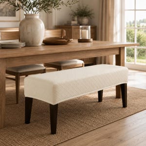 May include: A rectangular bench featuring a cream-colored upholstered seat with a subtle diagonal quilted pattern. The bench is supported by four straight, dark brown wooden legs that provide a stable base for the cushioned top surface.
