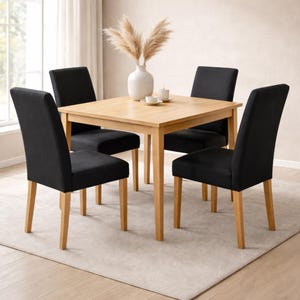 May include: A square wooden dining table with four black upholstered chairs. The table is light brown with a natural wood grain. The chairs have light brown wooden legs and a black seat and back. A white vase with dried plants sits on the table.