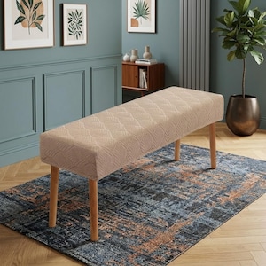 May include: A rectangular bench featuring a light beige, textured fabric cover with a repeating leaf-like pattern. The bench is supported by four cylindrical, light-colored wooden legs that extend outward at a slight angle from the base.