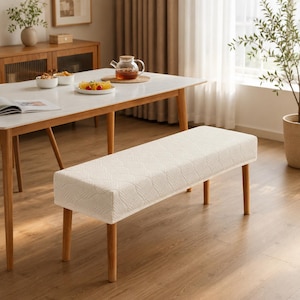 May include: A rectangular dining bench featuring a cream-colored, textured fabric cover with a subtle leaf-like pattern. The bench is supported by four light brown wooden legs, providing a stable base for seating in a dining room setting.