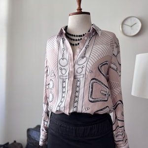 Może przedstawiać: Koszula zapinana na guziki z różowo-białym wzorem z czarnymi łańcuchami i geometrycznymi wzorami. Koszula z długim rękawem jest połączona z czarną spódnicą i czarnym naszyjnikiem z koralików. Koszula jest prezentowana na manekinie.