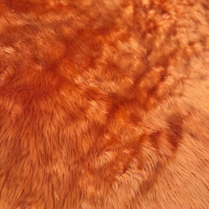 May include: Close-up of a textured, orange faux fur fabric. The fur has long, flowing fibers, creating a soft, plush appearance. The color is a warm, inviting shade of orange.