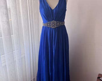 May include: A royal blue formal gown with a V-neckline and a flowing skirt. The dress features embellished shoulder straps and a jeweled belt at the waist. The dress is displayed on a mannequin.
