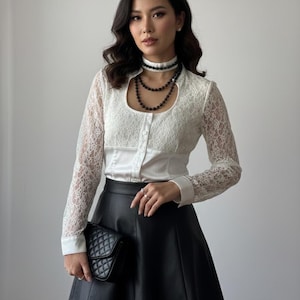 Könnte beinhalten: Eine weiße Spitzenbluse mit langen Ärmeln und Schlüsselloch-Ausschnitt, kombiniert mit einem schwarzen Lederrock und einer schwarzen Stepp-Clutch. Das Outfit wird durch eine schwarze Perlenkette ergänzt.