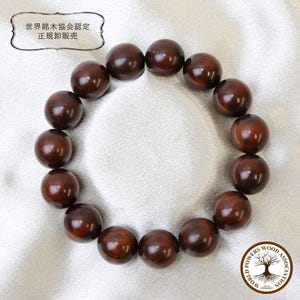 Op de afbeelding: Een cirkelvormige armband bestaande uit vijftien gepolijste, donkerbruine houten kralen. De afbeelding bevat tekst in Japanse karakters binnen een decoratieve rand en een rond logo met een boomafbeelding en de tekst World Powers Wood Association.