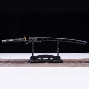 May include: A black katana sword with a curved blade and a black handle wrapped in a diamond pattern. The sword features a gold-colored tsuba and a black and white cord tied around the scabbard, resting on a black wooden display stand.