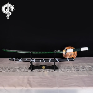 Könnte beinhalten: Ein japanisches Katana-Schwert mit grüner Klinge und einer schwarz-weiß gemusterten Scheide. Das Schwert wird auf einem schwarzen Ständer mit goldenem Emblem präsentiert. Der Griff ist weiß und grün.
